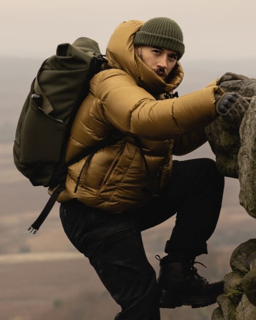 Man rock climbing wearing a gold Canada Goose puffer jacket, green beanie, black pants and green backpack