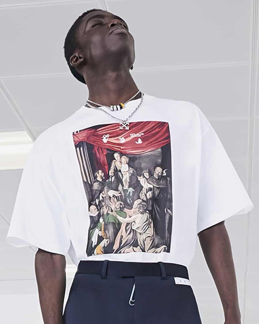 Black man wearing a white large graphic print T-shirt by Off-White with blue pants