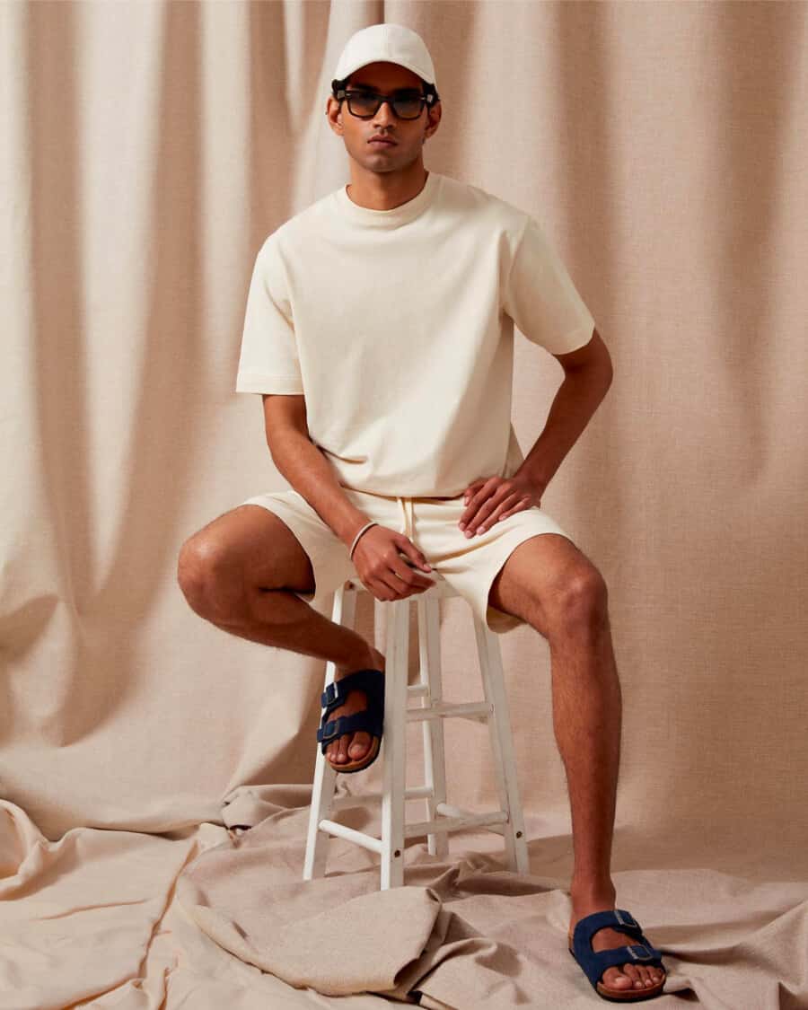 Man wearing an oversized cream T-shirt with matching cream shorts, sunglasses, baseball cap and slide sandals