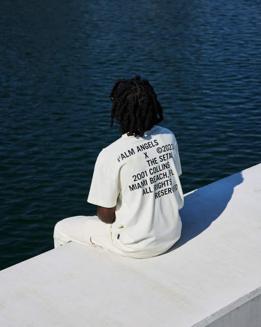 Black man wearing a white printed back Palm Angels T-shirt with white pants.