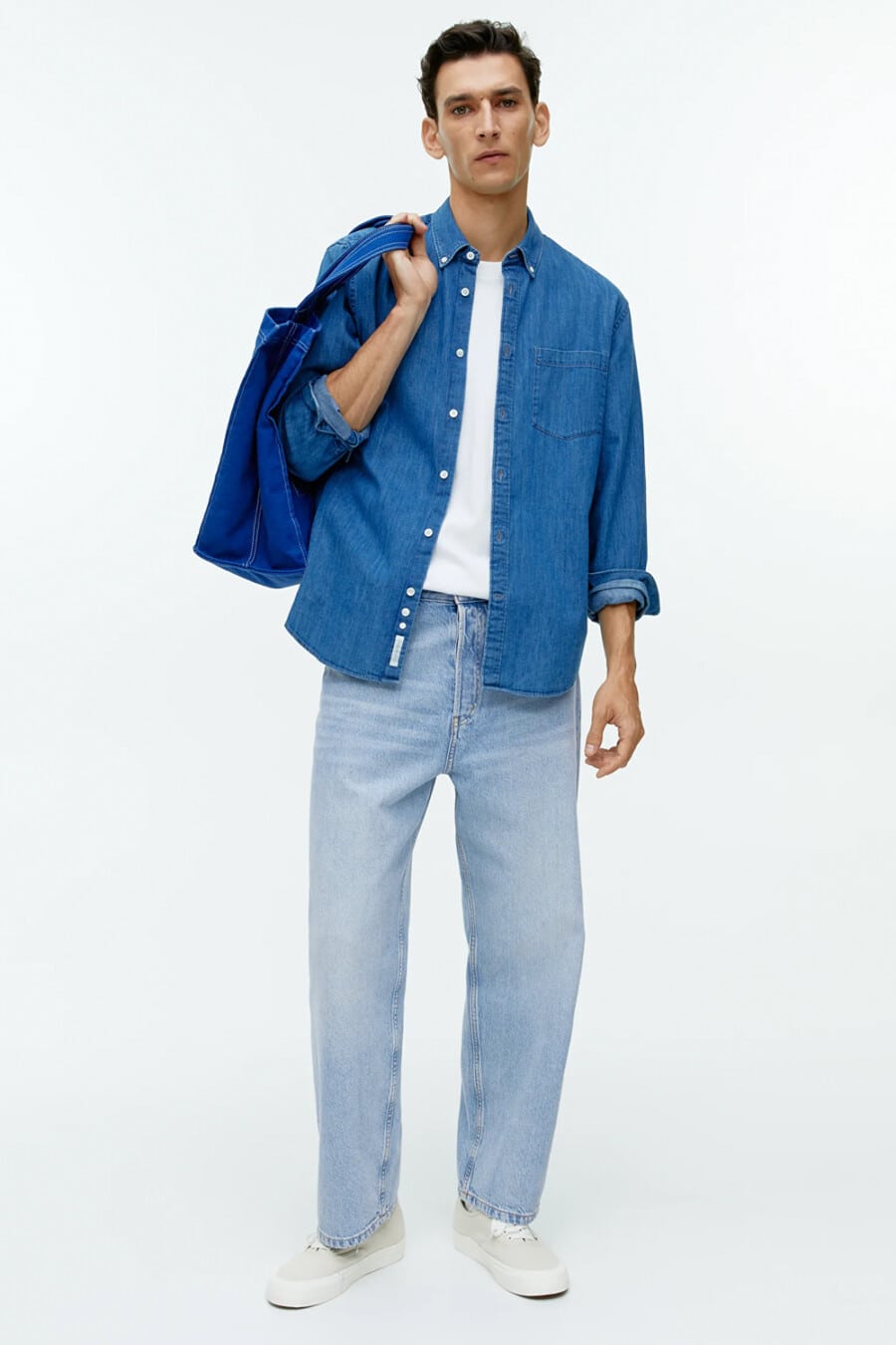 Men's baggy light wash jeans, white T-shirt, open blue denim shirt, blue tote bag and off-white canvas skate shoes outfit