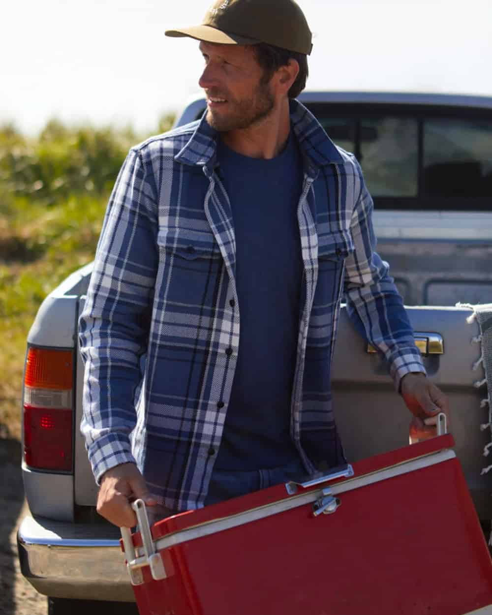 Man wearing Outerknown blue T-shirt, mid-wash blue jeans, blue/white flannel check overshirt and khaki baseball cap carrying a cooler