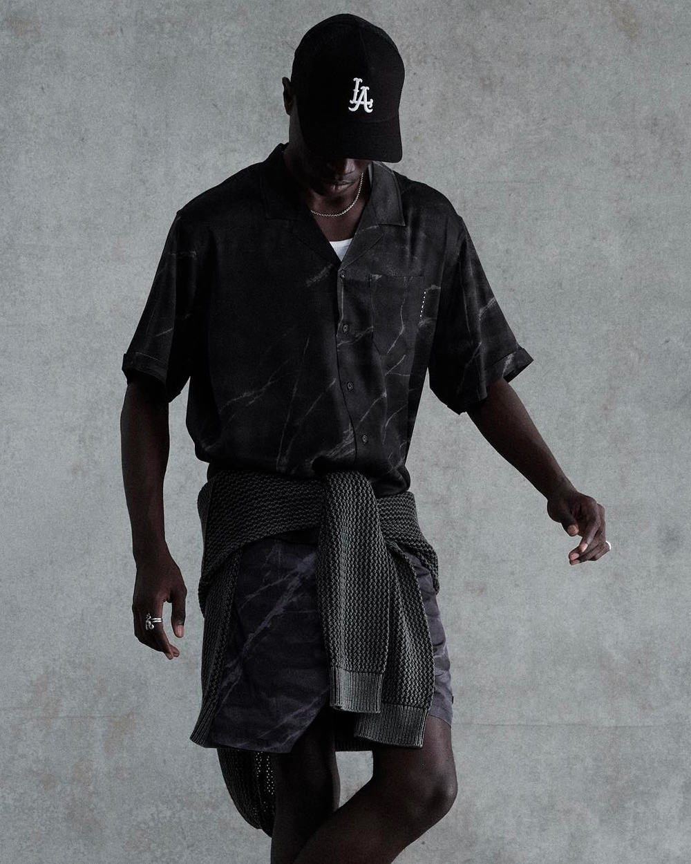 Man wearing black matching shirt and shorts, black LA baseball cap and tied grey sweater around the waist by Stampd