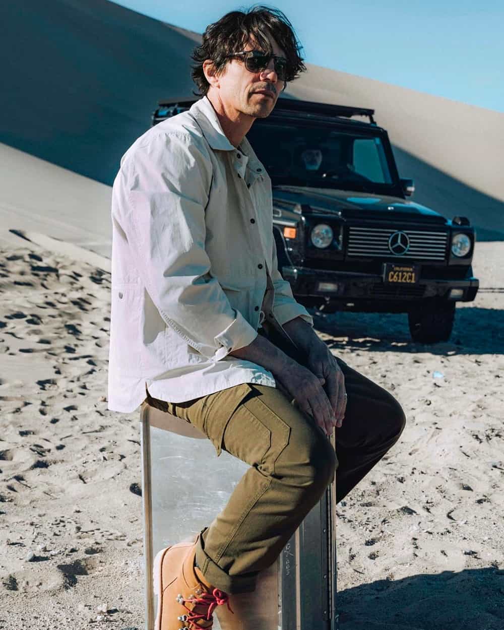 Man wearing AETHER Apparel khaki cargo pants, tan hiking boots and white overshirt with black sunglasses