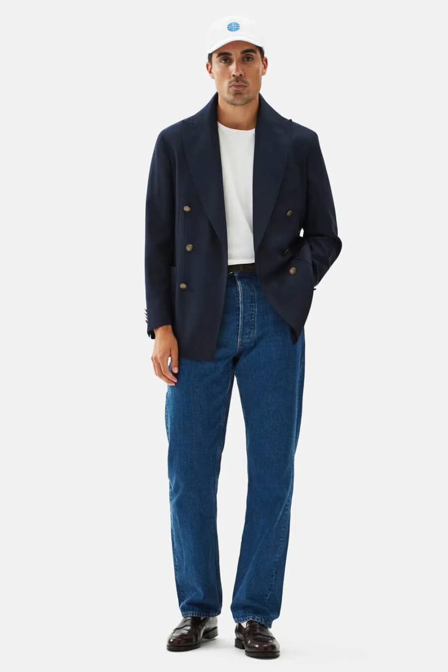 Men's navy double-breasted blazer, mid-wash jeans, white T-shirt tucked in, white baseball cap and brown leather penny loafers outfit
