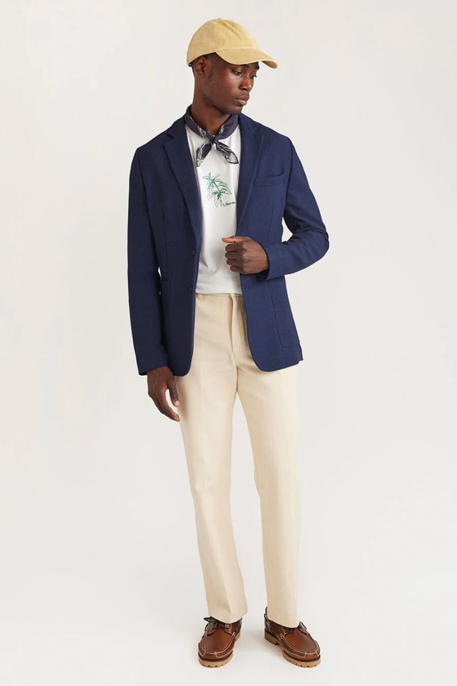 Navy single-breasted blazer, light cream chinos, white printed T-shirt, navy neckerchief, beige baseball cap and brown leather chunky boat shoes outfit