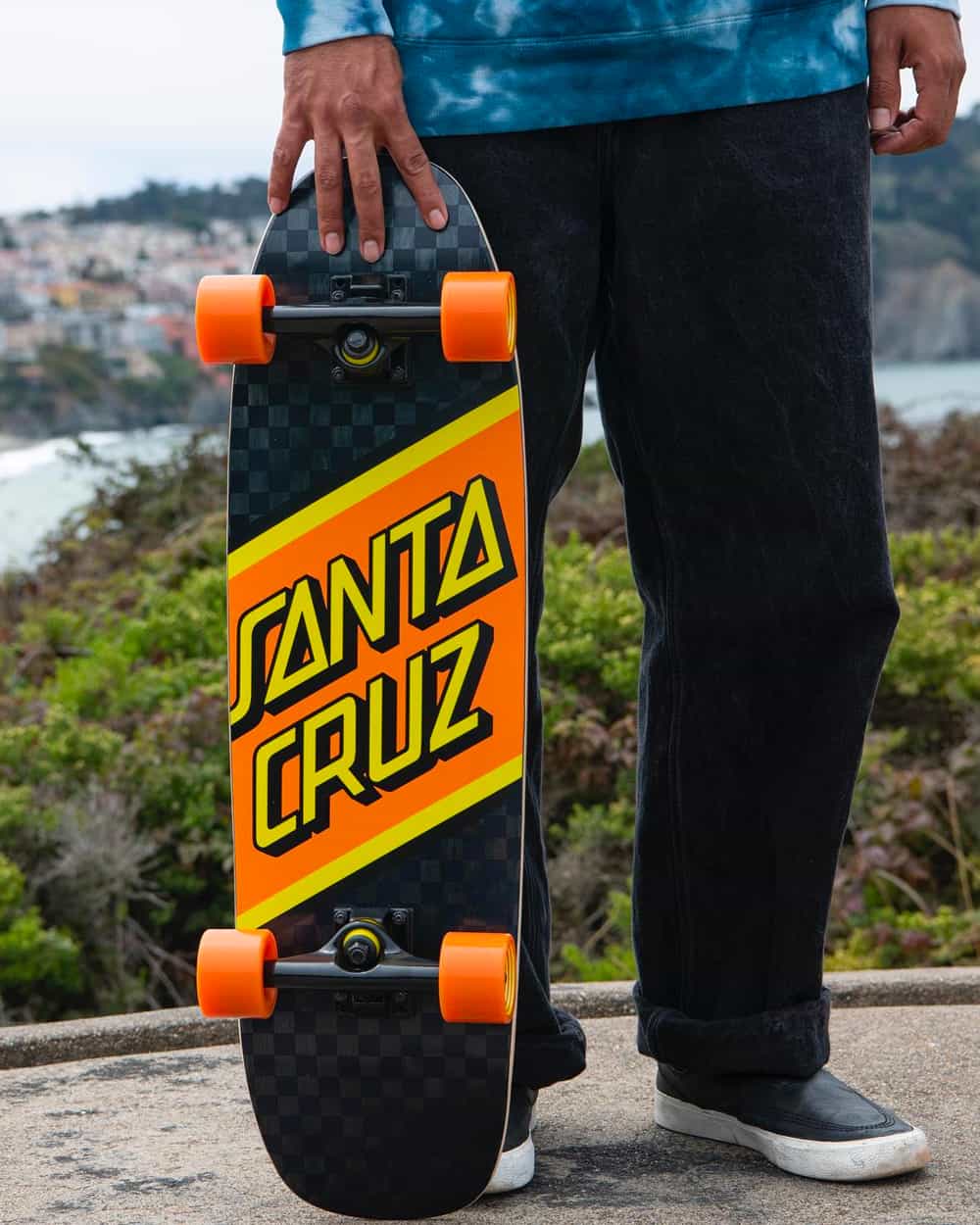 Man wearing baggy black jeans and blue tie dye top holding a branded Santa Cruz skateboard