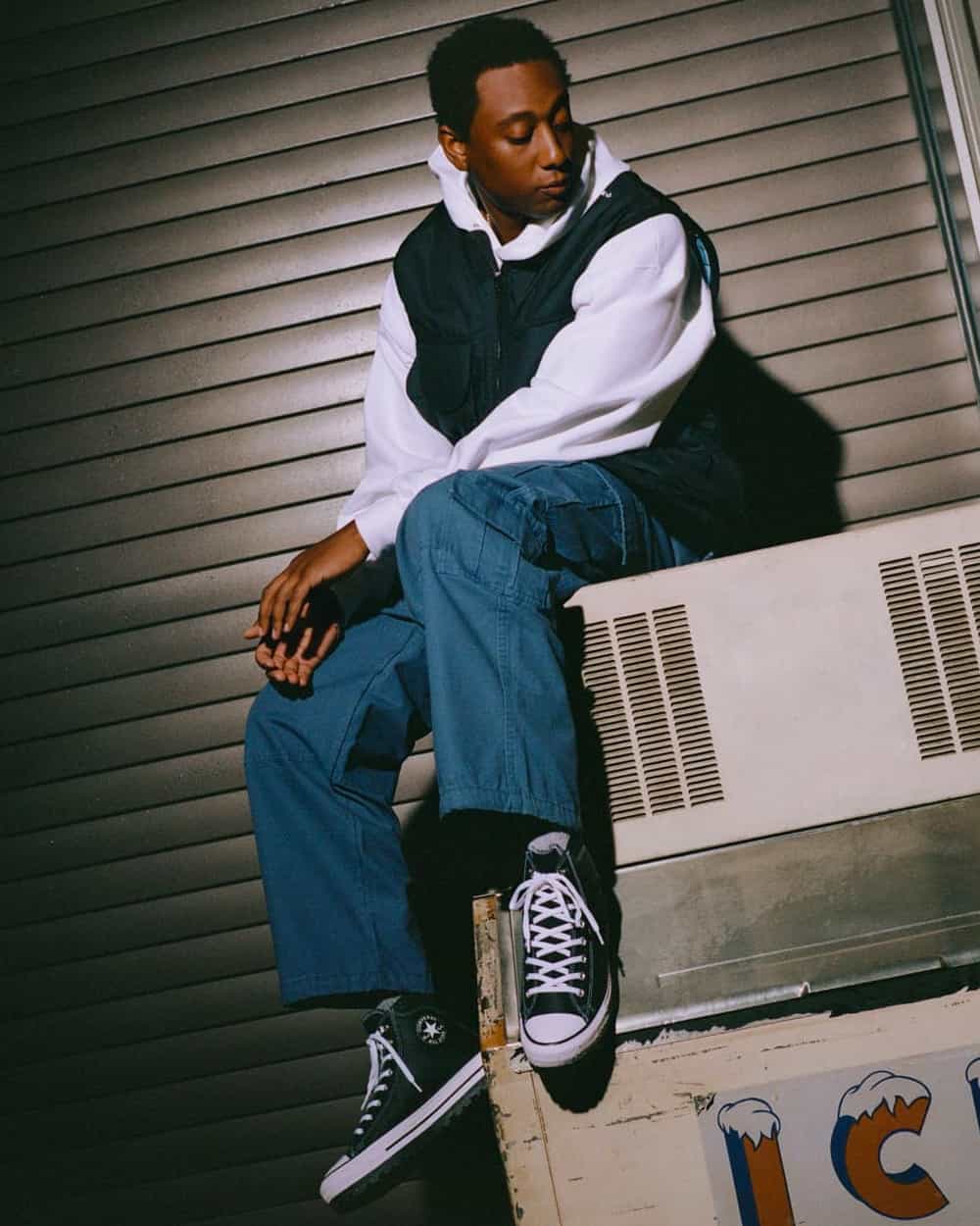 Man wearing white hoodie, black vest, loose blue canvas pants and Converse black high-tops