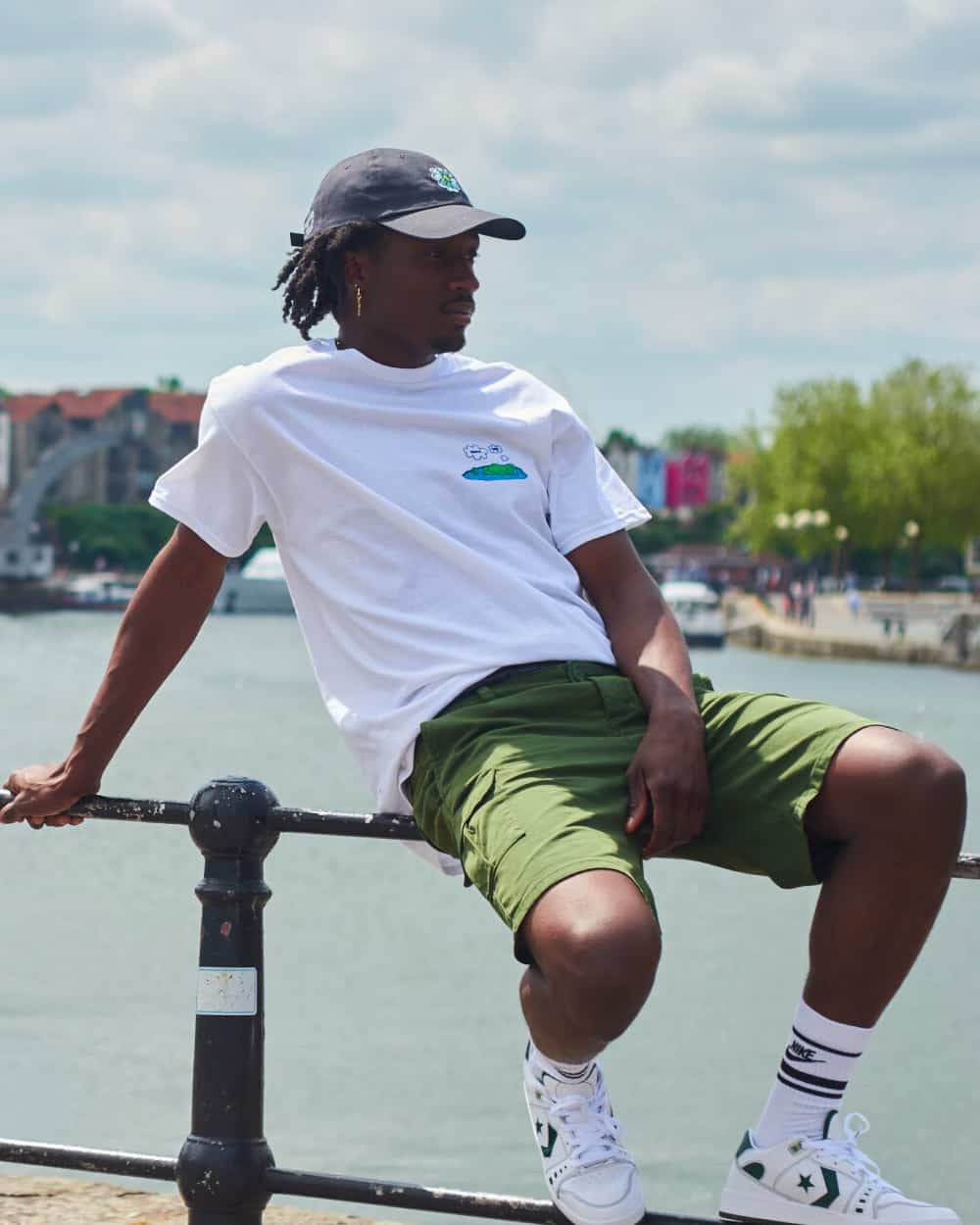 Black man wearing Route One green shorts, white printed T-shirt, black baseball cap and Converse One Star skate shoes with white Nike tube socks.