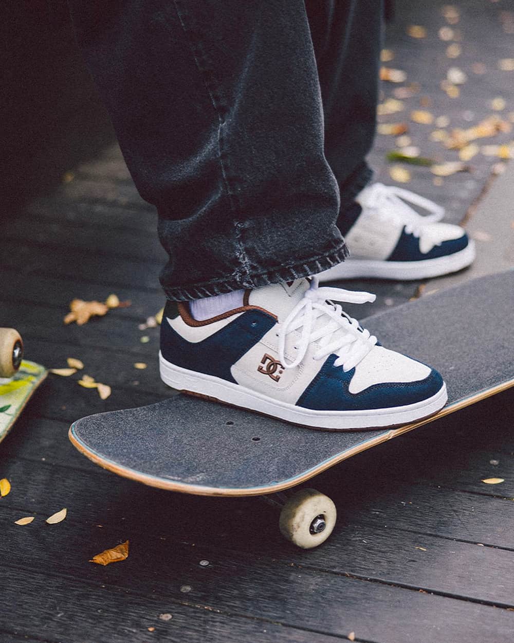 A pair of DC skate shoes worn on feet with white socks and loose black jeans with a skateboard