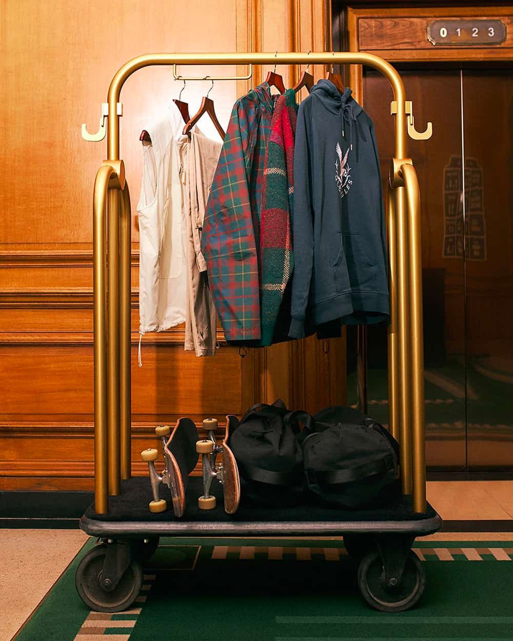 A hotel porter hanging rail with multiple Pop Trading Company skatewear pieces hanging on it, along with two skateboards