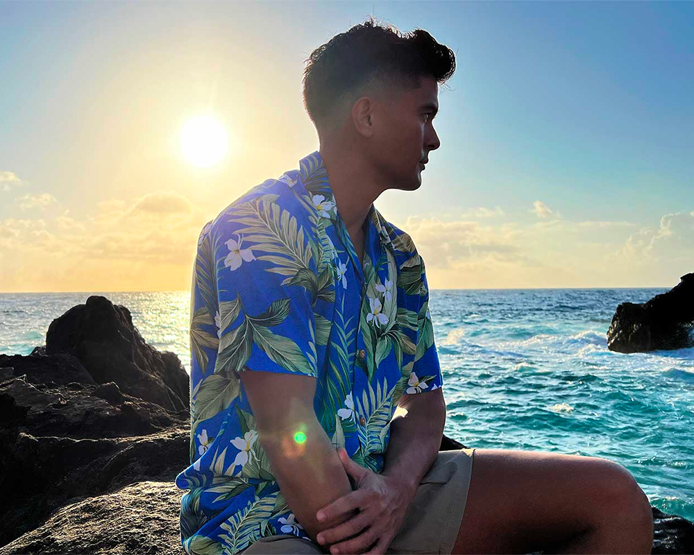 Man wearing a blue palm print original Paradise Found Hawaiian shirt outside by the sea