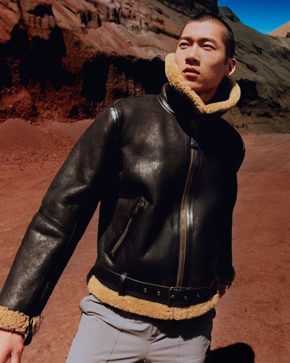 Asian man wearing a brown funnel neck shearling jacket with grey pants