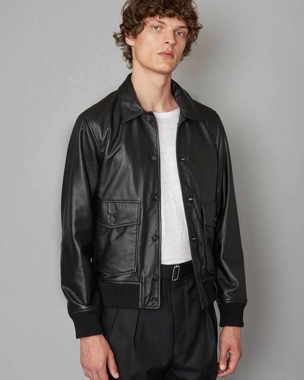 Man wearing Officine Générale black pleated pants, tucked in white T-shirt and black leather collared flight jacket