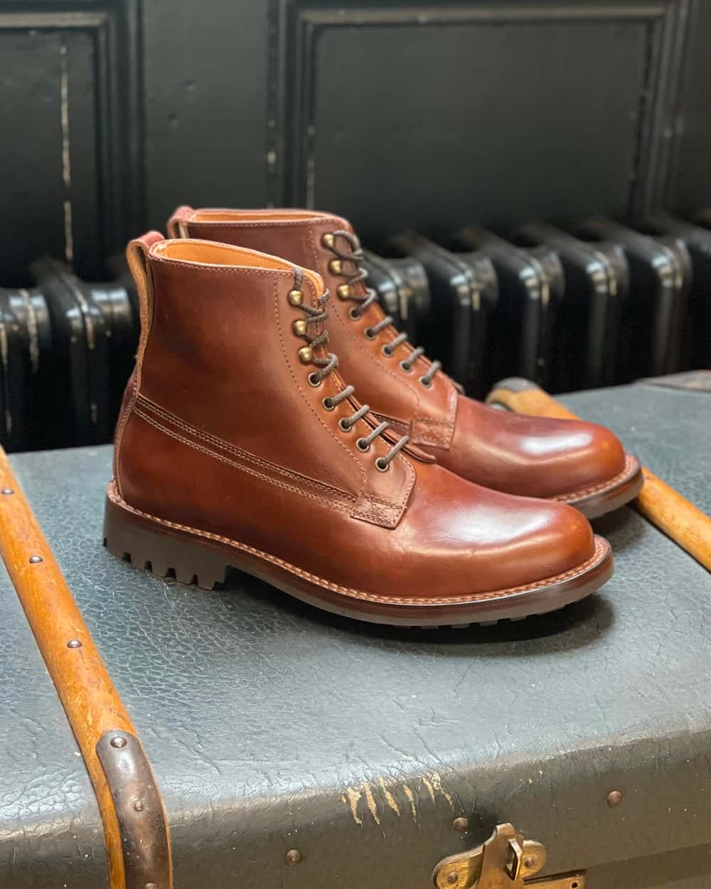 A pair of brown leather Grenson men's military Derby boots with lugged sole sitting on top of a chest