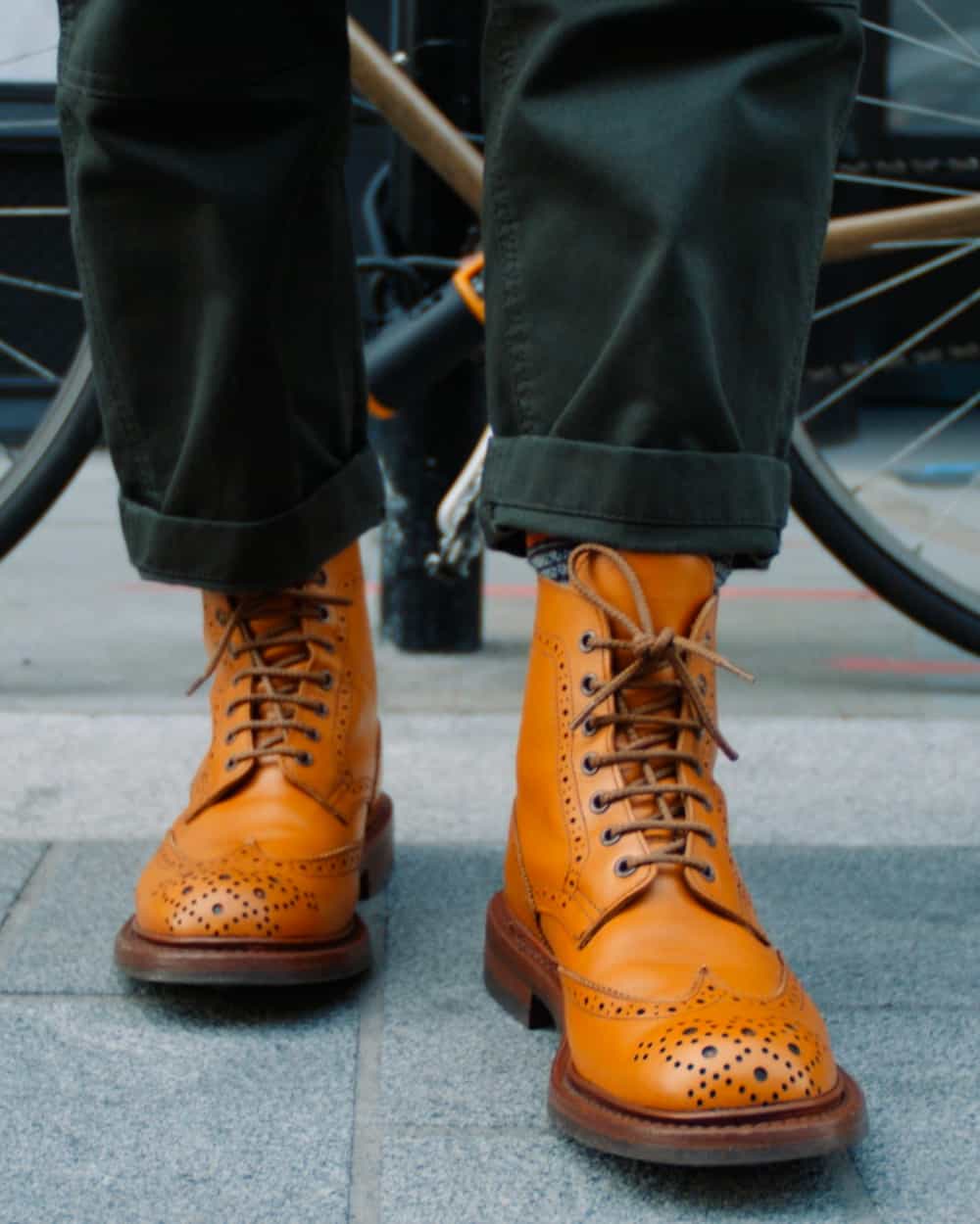 A pair of men's tan leather Tricker's brogue boots worn on feet with turned up blue-green pants