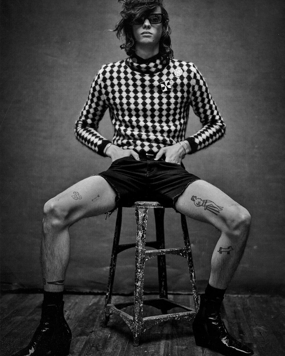 Man sitting on stool wearing black/white checkerboard turtleneck, short black shorts and black leather Chelsea boots all by Saint Laurent