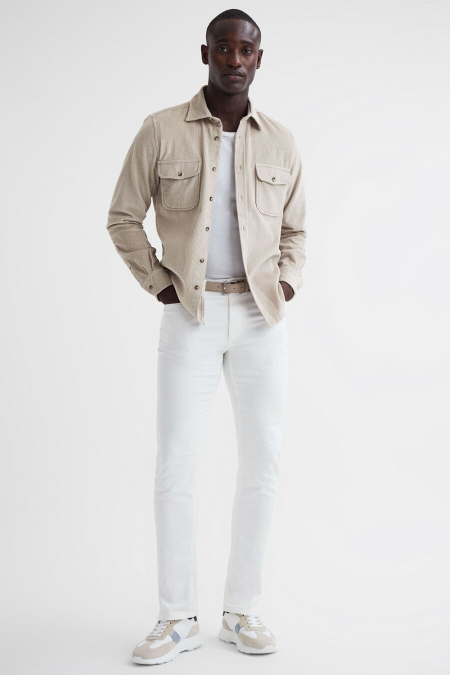 Men's white jeans, white T-shirt tucked in, beige corduroy shacket, beige suede belt and white/beige sneakers outfit