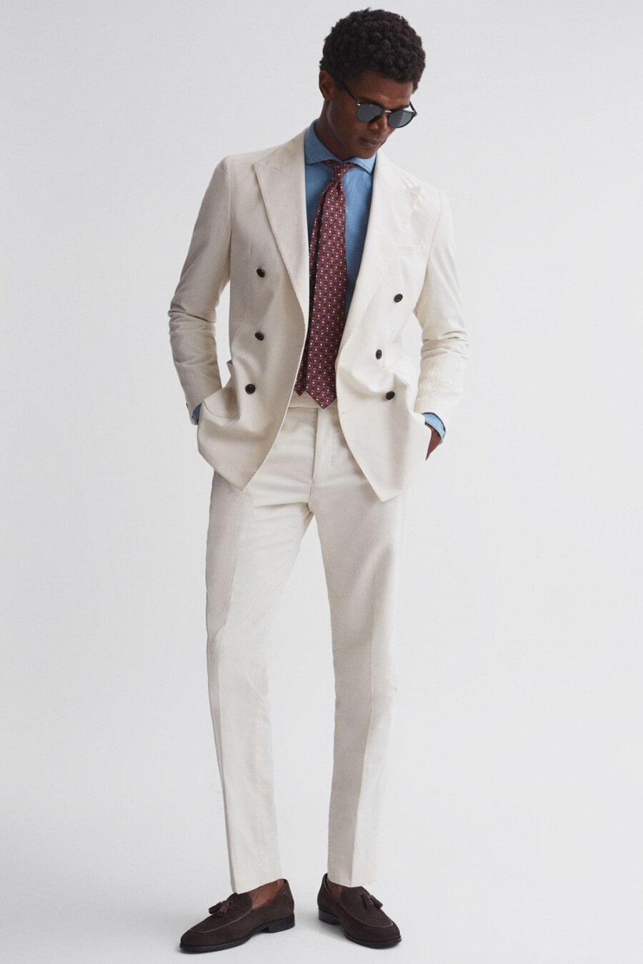 Men's cream double-breasted suit, chambray shirt, burgundy patterned tie and brown suede tassel loafers worn sockless outfit