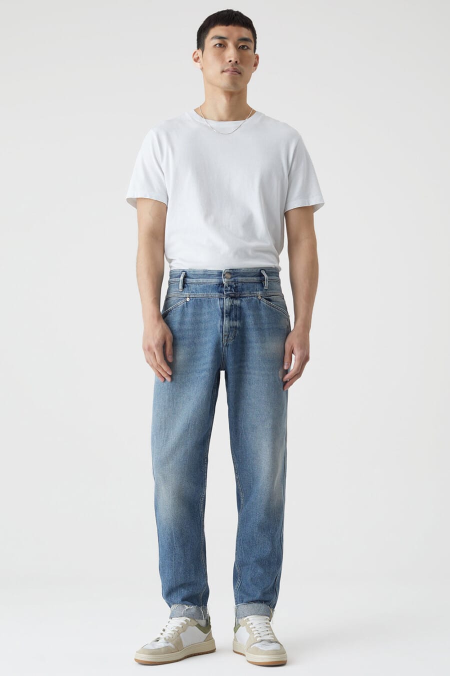 Men's baggy light wash jeans, tucked in white T-shirt, silver necklace chain and white/beige chunky sneakers