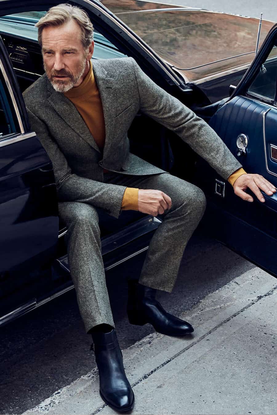 Man wearing grey flannel textured suit, long-sleeve mustard top and black leather Chelsea boots sitting in vintage car