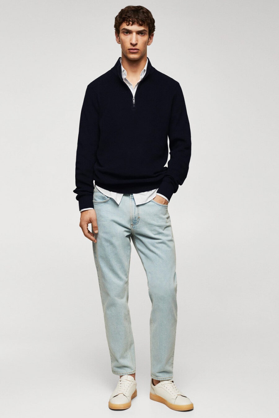 Men's light wash blue jeans, white shirt, navy half-zip sweater and white gum sole sneakers outfit