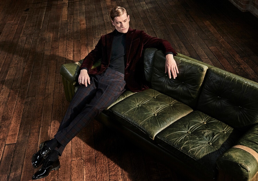 Man wearing check pants, black turtleneck, velvet burgundy blazer and black patent Oxford shoes sitting on green leather sofa