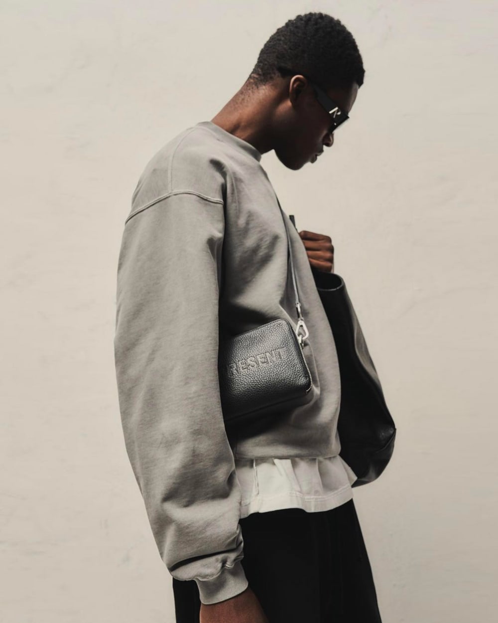 Man wearing oversized grey Represent sweatshirt over long white T-shirt with black pants and a crossbody bag