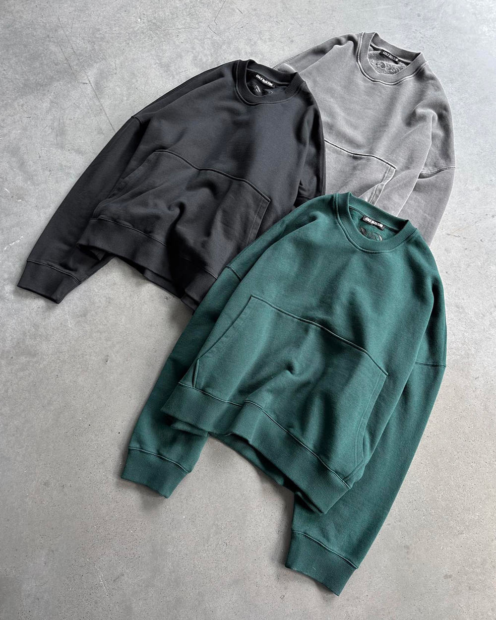 Three oversized sweatshirts by Cole Buxton in green, charcoal and grey laid out on concrete floor