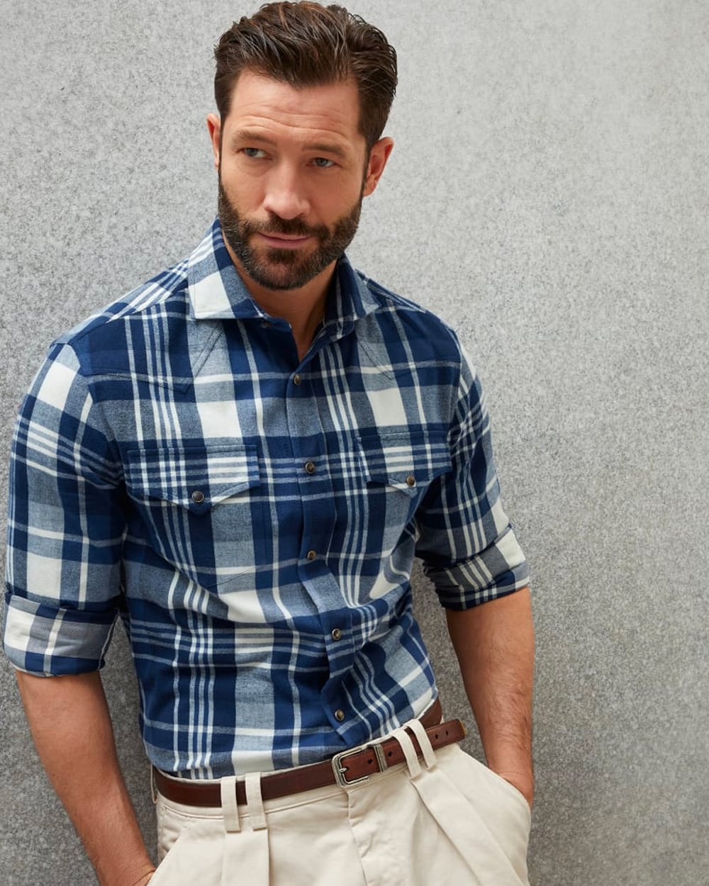 Man wearing blue/white check flannel shirt by Brunello Cucinelli tucked into off-white pleated pants with a brown leather belt
