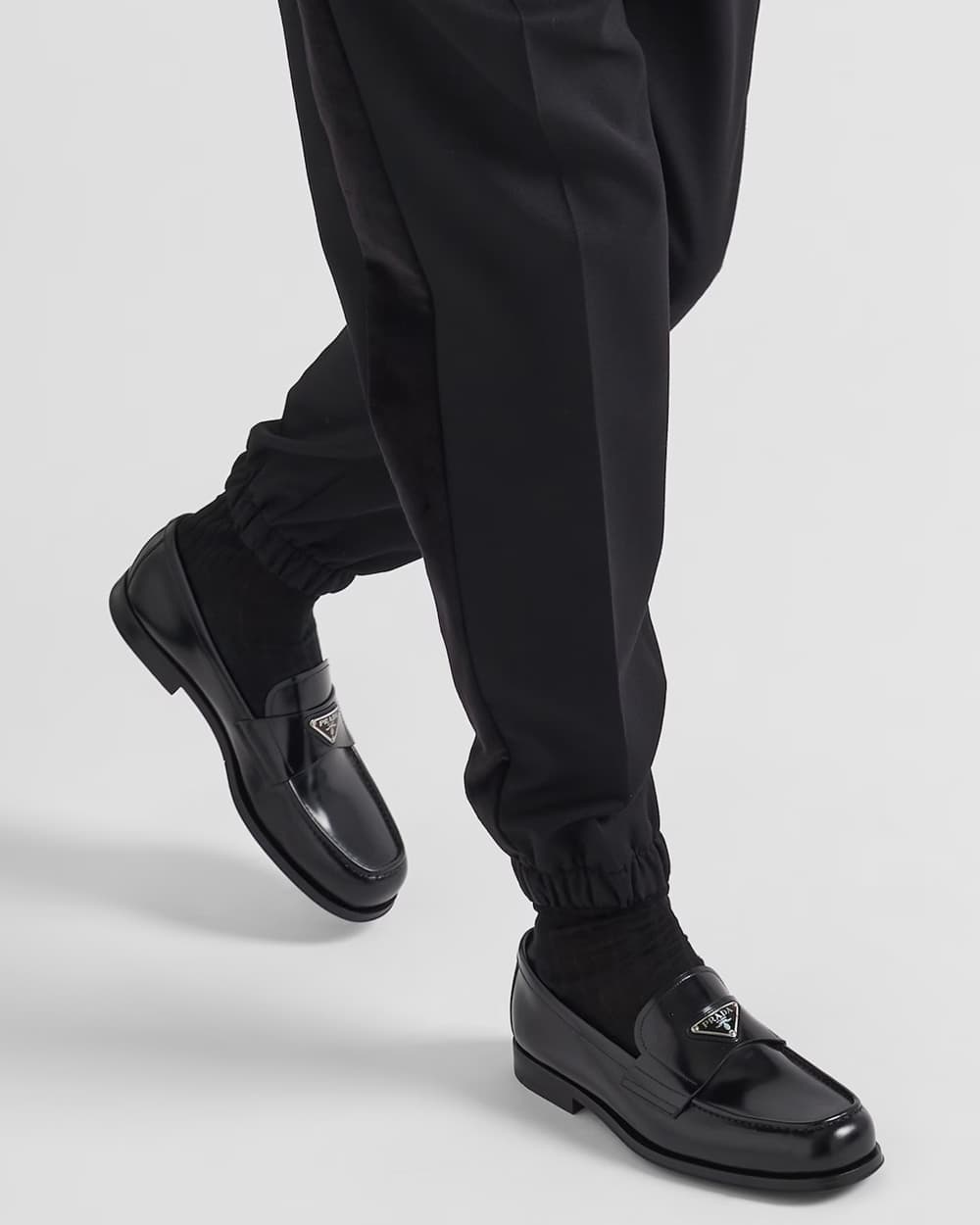 Man wearing black Prada leather loafers with silver logo adorned with black tailored cuffed pants and black socks