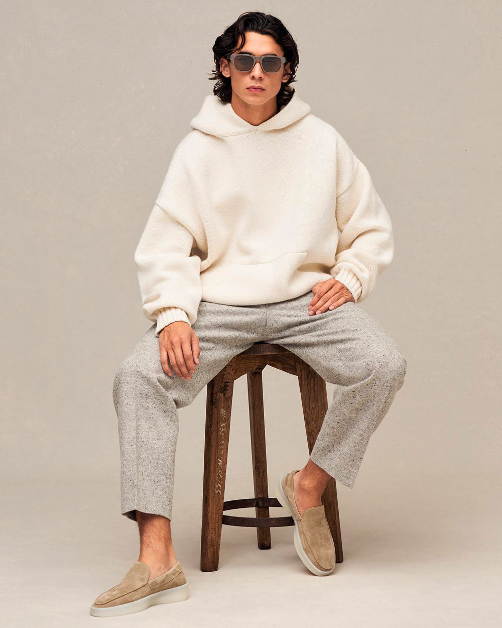 Man sitting on stool wearing oversized cream hoodie, cropped light grey pants and taupe suede loafers by Fear of God