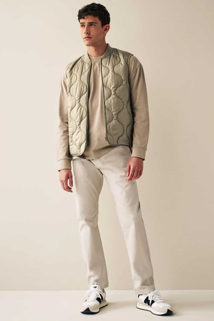 Men's off-white chinos, beige long-sleeve top, light green padded vest and white New Balance sneakers outfit