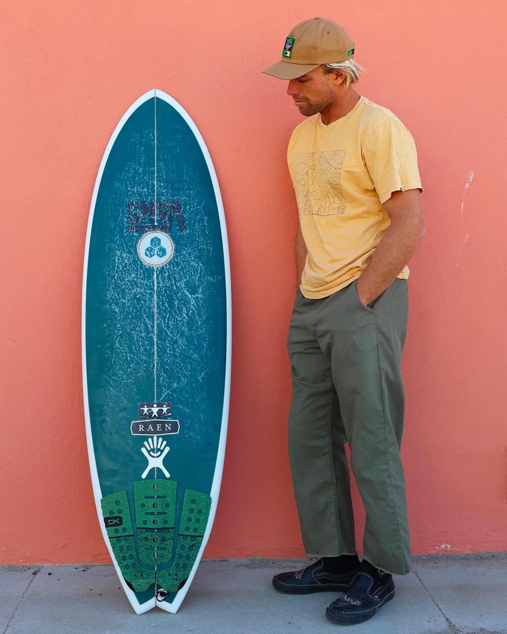Man wearing cropped green pants, yellow T-shirt and beige baseball cap with a blue Channel Islands Surfboard