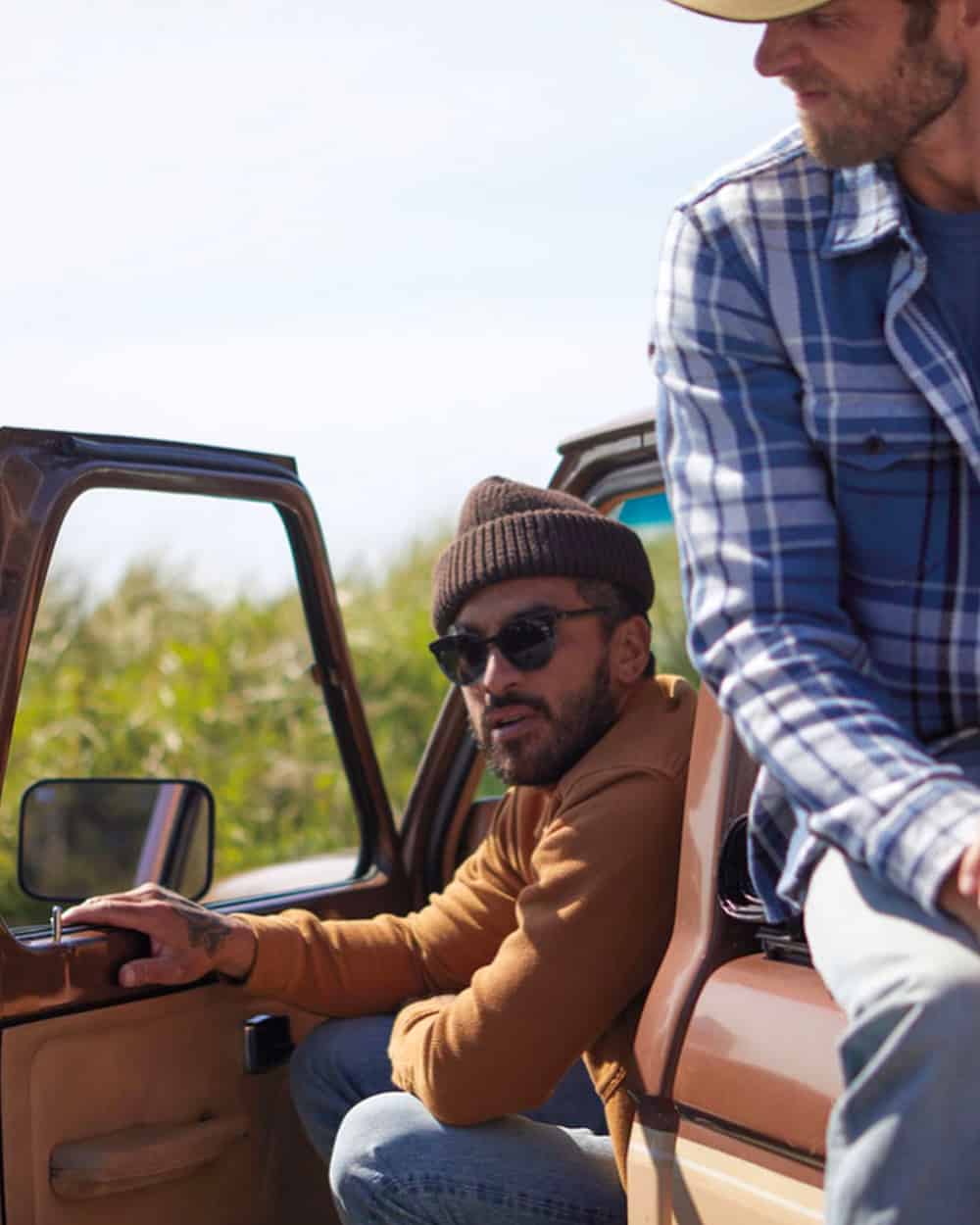 Man wearing Outerknown mustard top, brown beanie, round lens sunglasses and light wash jeans sitting in car