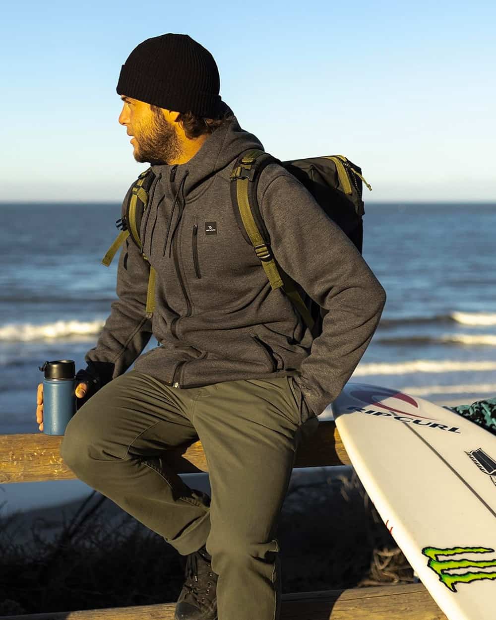 Man wearing Rip Curl green pants, grey fleece jacket and black beanie next to his surfboard