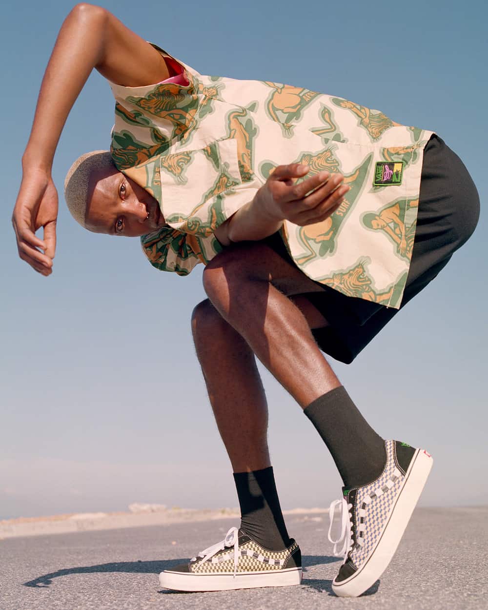 Black man wearing checkerboard Vans skate shoes, black shorts, black socks and cream, green orange printed short sleeve shirt