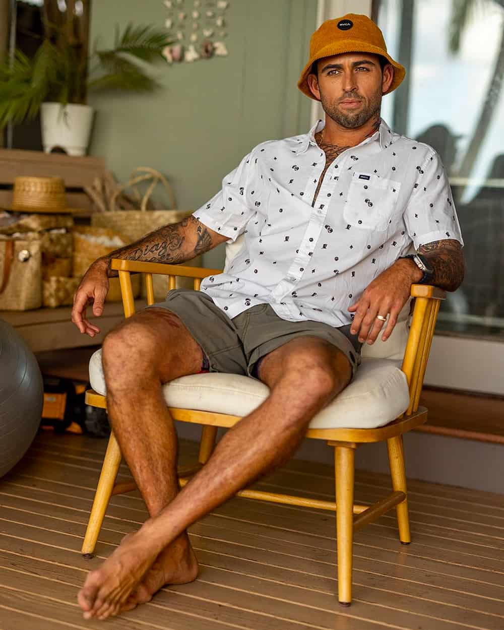 Man wearing RVCA grey shorts, printed white short sleeve shirt and orange bucket hat sitting on chair