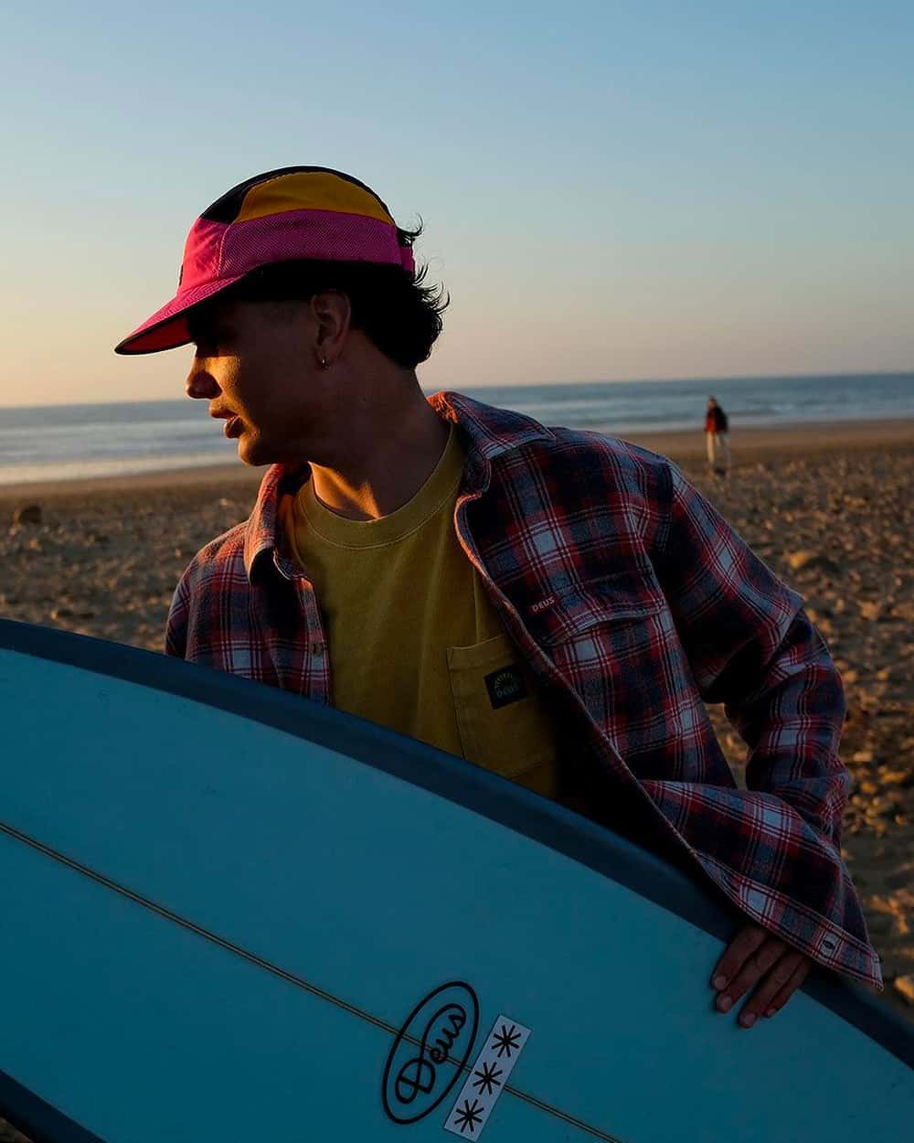 Man holding light blue surfboard wearing Deus Ex Machina red check flannel shirt, yellow T-shirt and multicolored cap