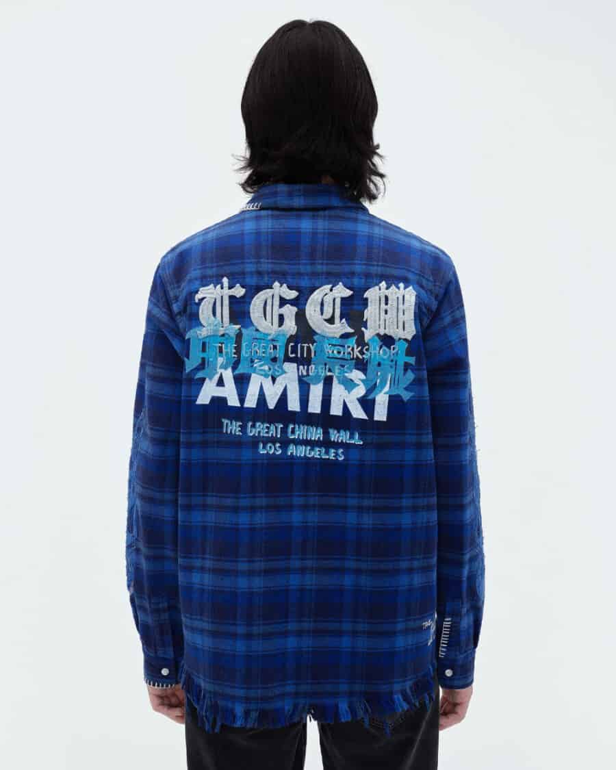 Reverse shot of man wearing a blue flannel checked shirt by AMIRI with printed branding to the back