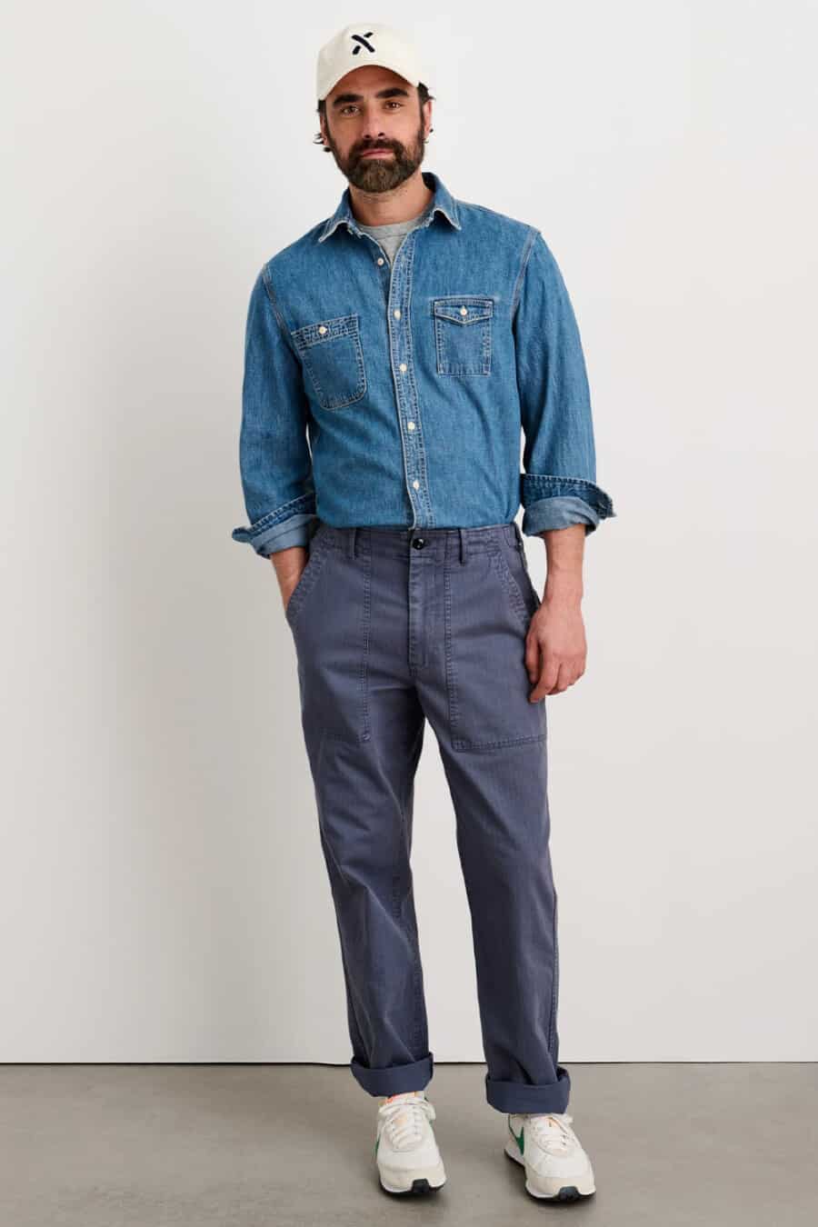 Men's grey-blue utility pants, tucked in grey T-shirt, mid-blue denim shirt with rolled sleeves, white baseball cap and white Nike sneakers outfit
