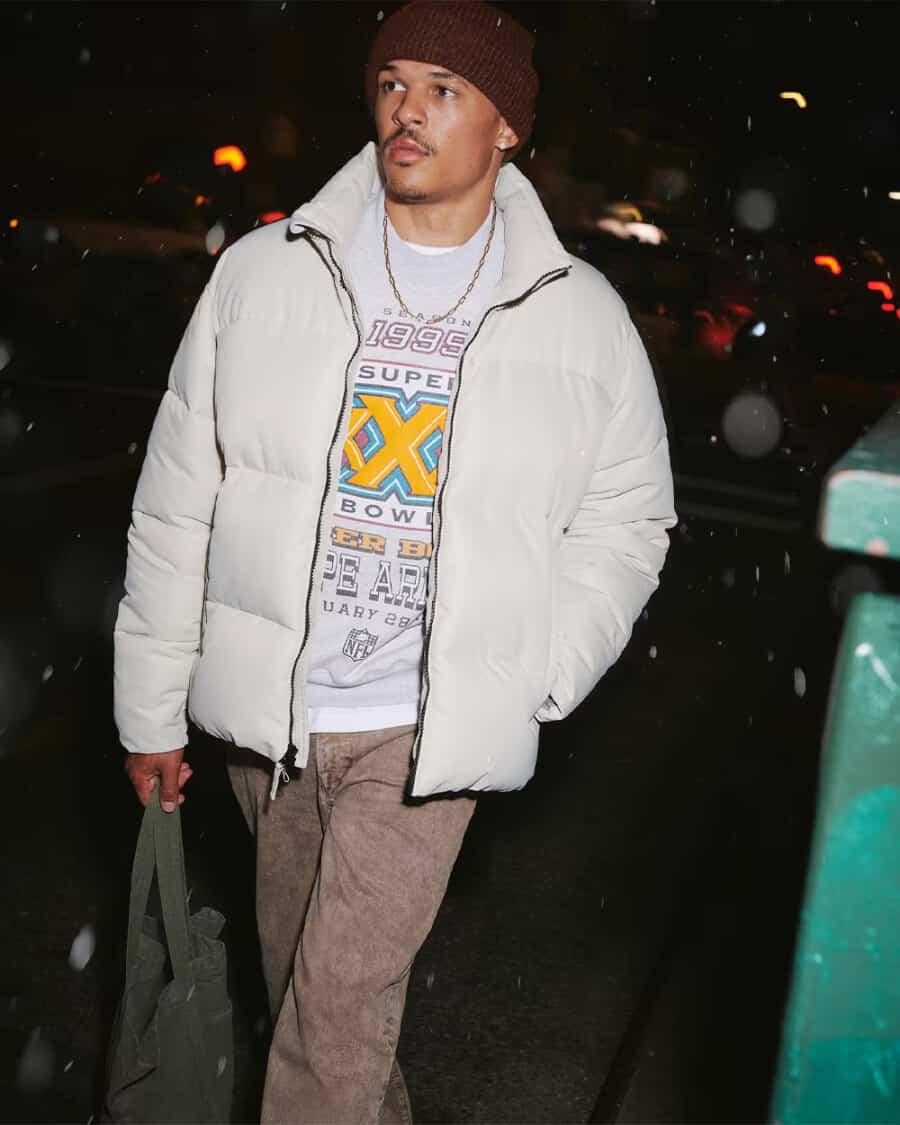 Man wearing white puffer jacket over a printed grey sweatshirt and brown pants with a brown beanie