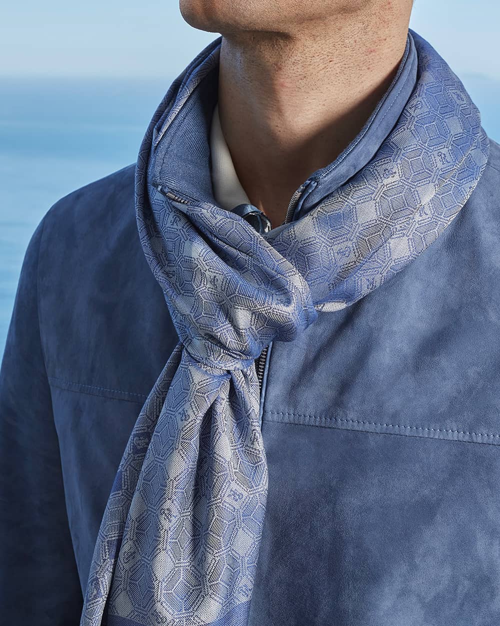 Close up of a man wearing a blue suede jacket with a blue patterned knotted Stefano Ricci scarf around his neck