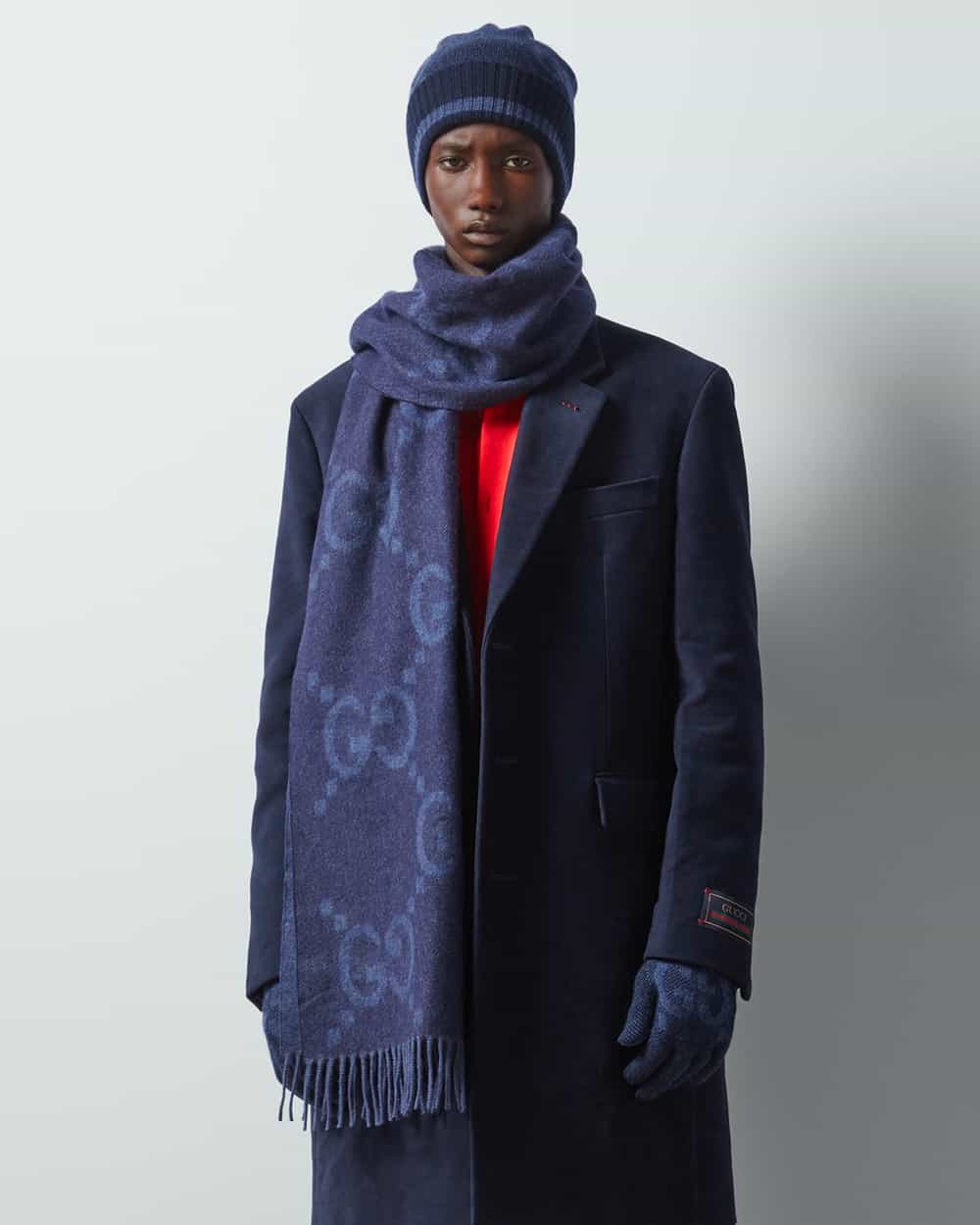 Man in bright red sweater and navy overcoat wearing a Gucci oversized logo scarf in navy