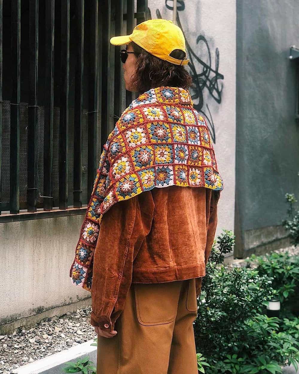 Man in brown pants, tan suede jacket and bright yellow baseball cap with Story MFG knitted flower pattern blanket scarf over shoulders