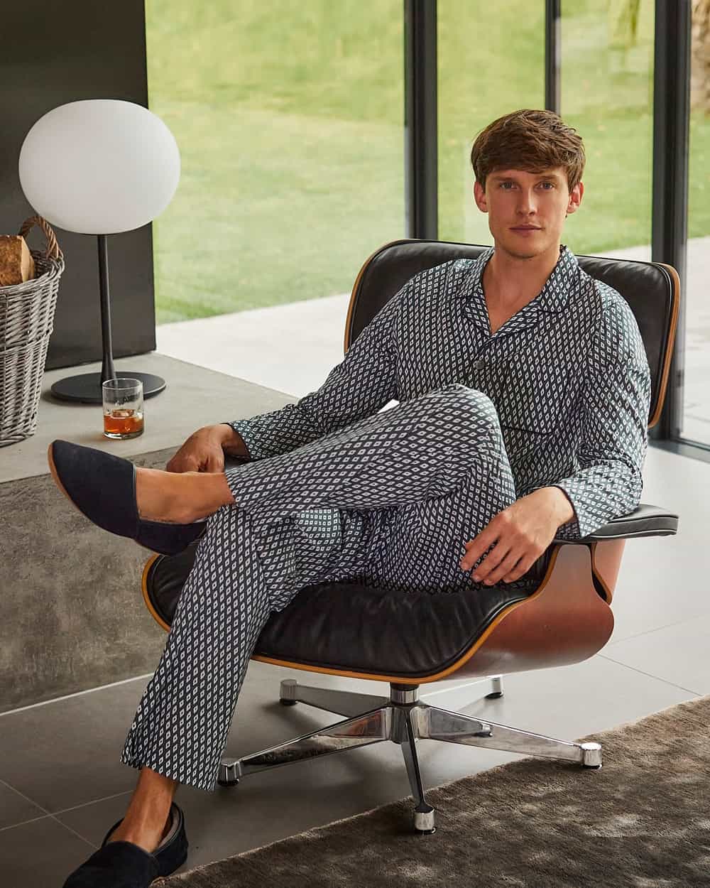 Man lounging in eames chair in luxury printed navy/white pajamas and navy slippers by Derek Rose