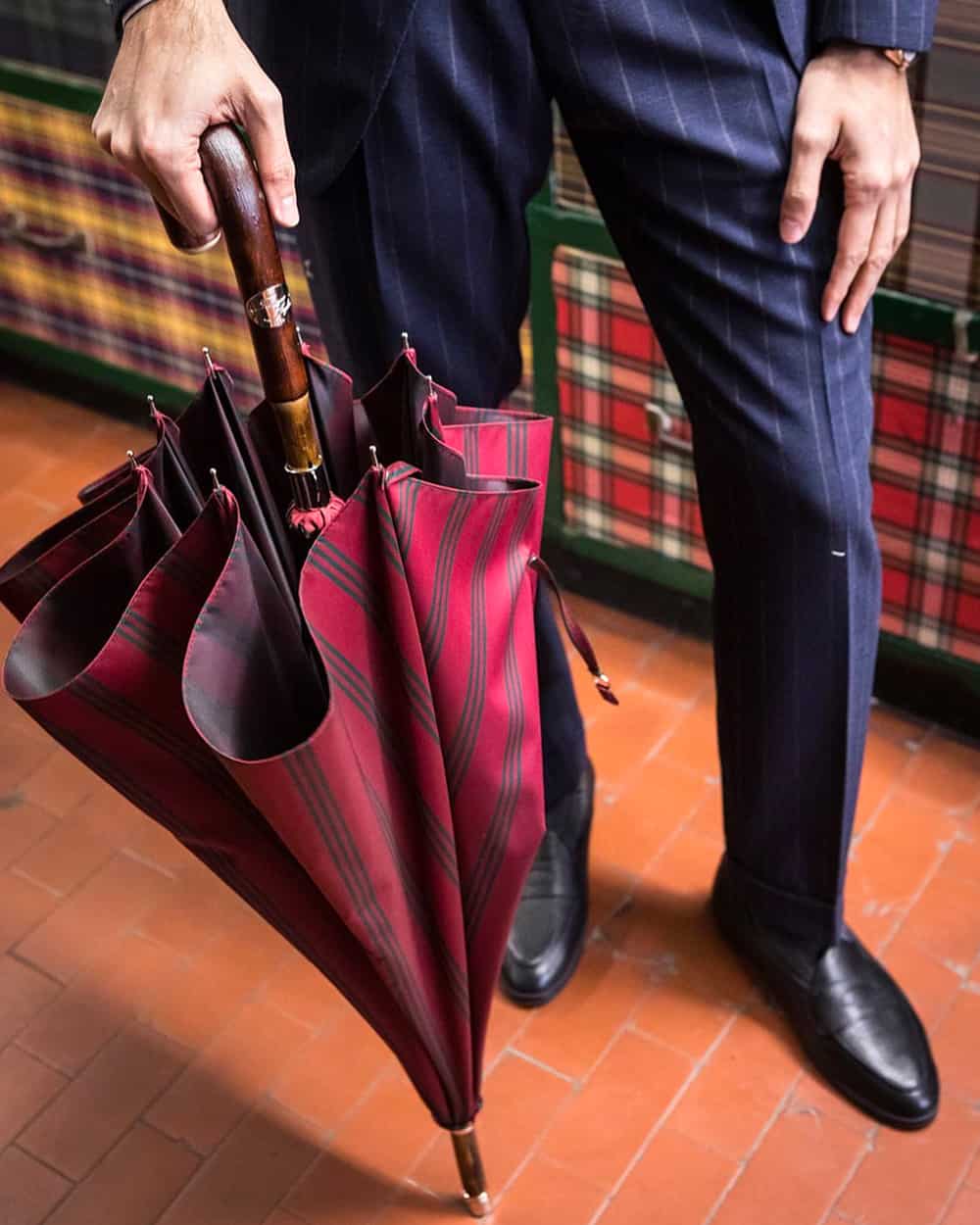Man in a navy pinstripe suit holding a wooden handle Francesco Maglia umbrella with a burgundy stripe canopy