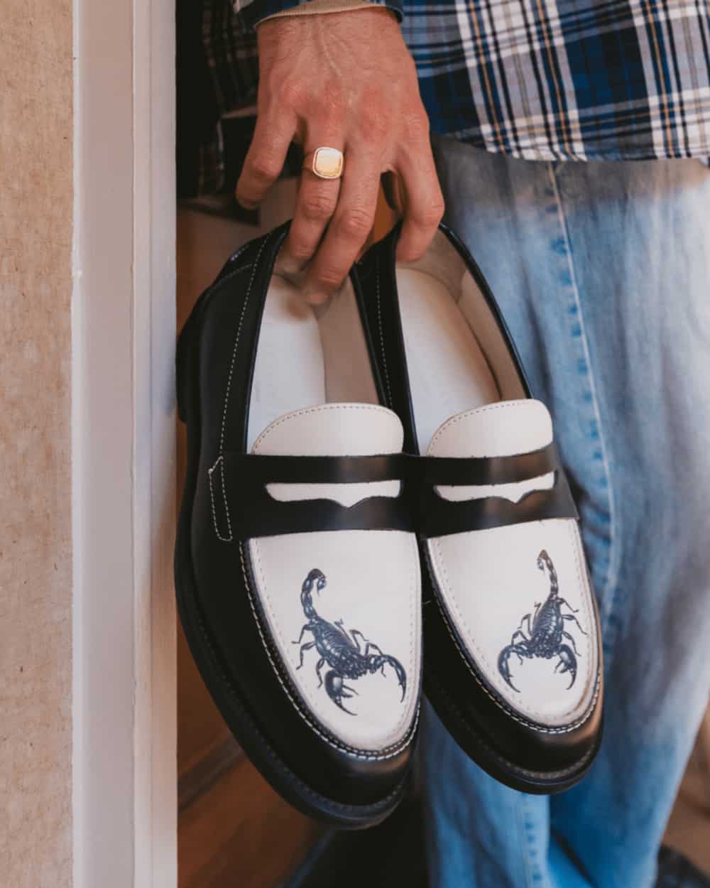 Man holding a pair of black and white spectator penny loafers with a blue printed scorpion on the toe by Duke & Dexter