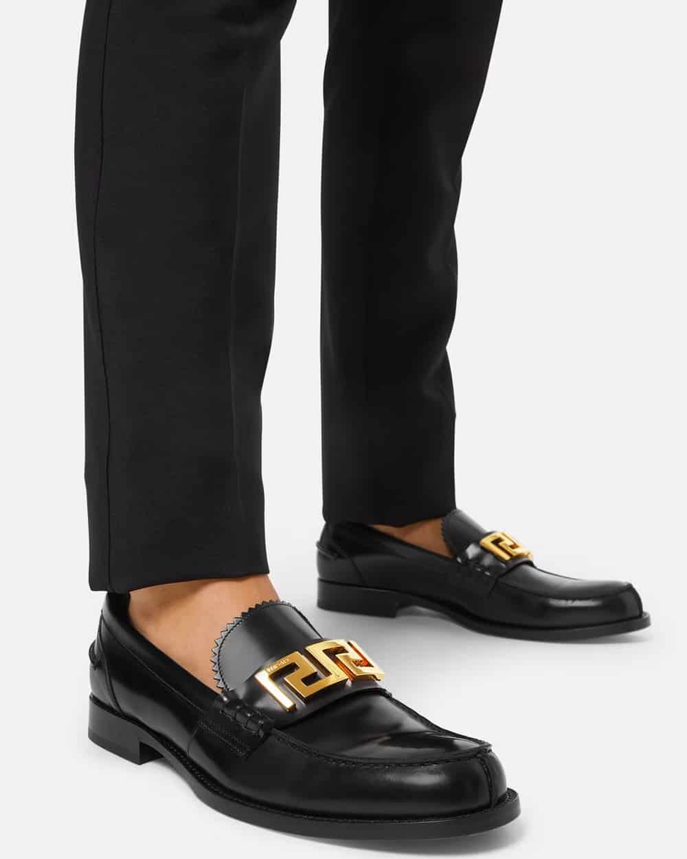 Man wearing black leather Versace loafers with gold metal branding sockless with black pants.