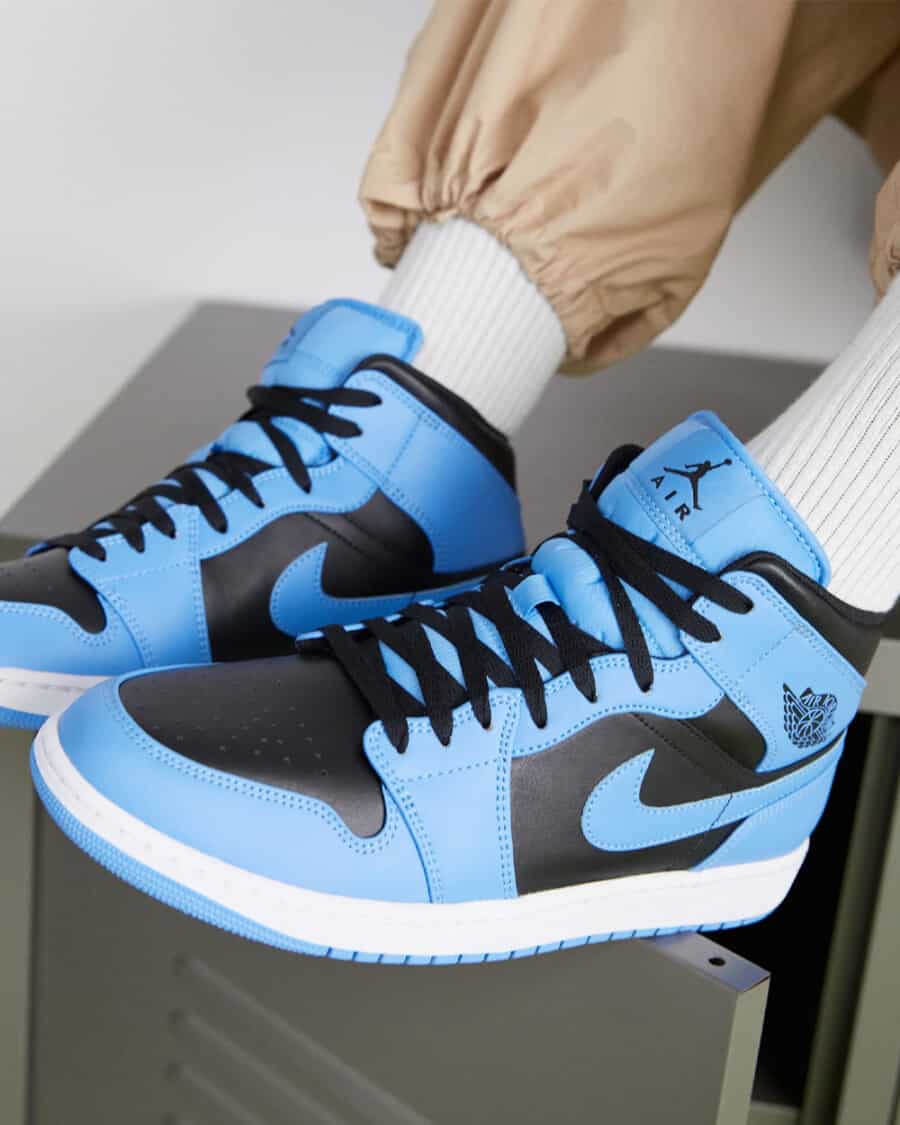 Pair of Air Jordan 1 sneakers in black and University blue worn on feet with white socks and khaki pants